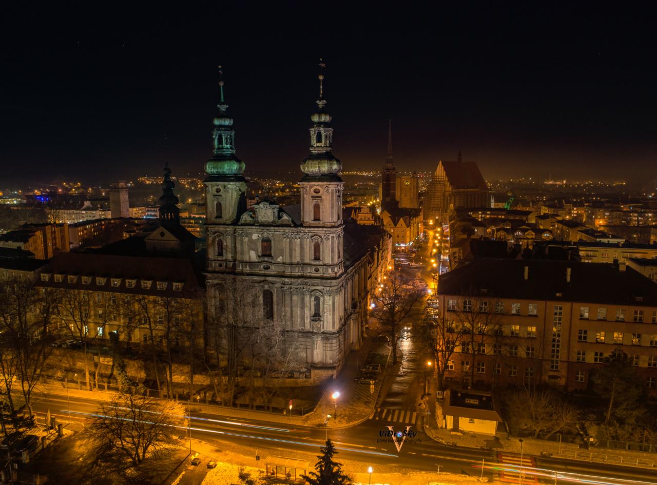 Nocny widok z lotu ptaka na oświetlone miasto z barokowym kościołem z dwiema wieżami na pierwszym planie, długie ekspozycje świateł samochodów na ulicach.
