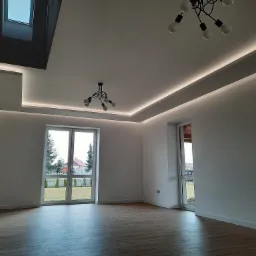 Jasne, przestronne wnętrze po remoncie z parkietem, oknami balkonowymi i nowoczesnym oświetleniem sufitowym, w tym listwy LED i lampy wiszące z odsłoniętymi żarówkami.