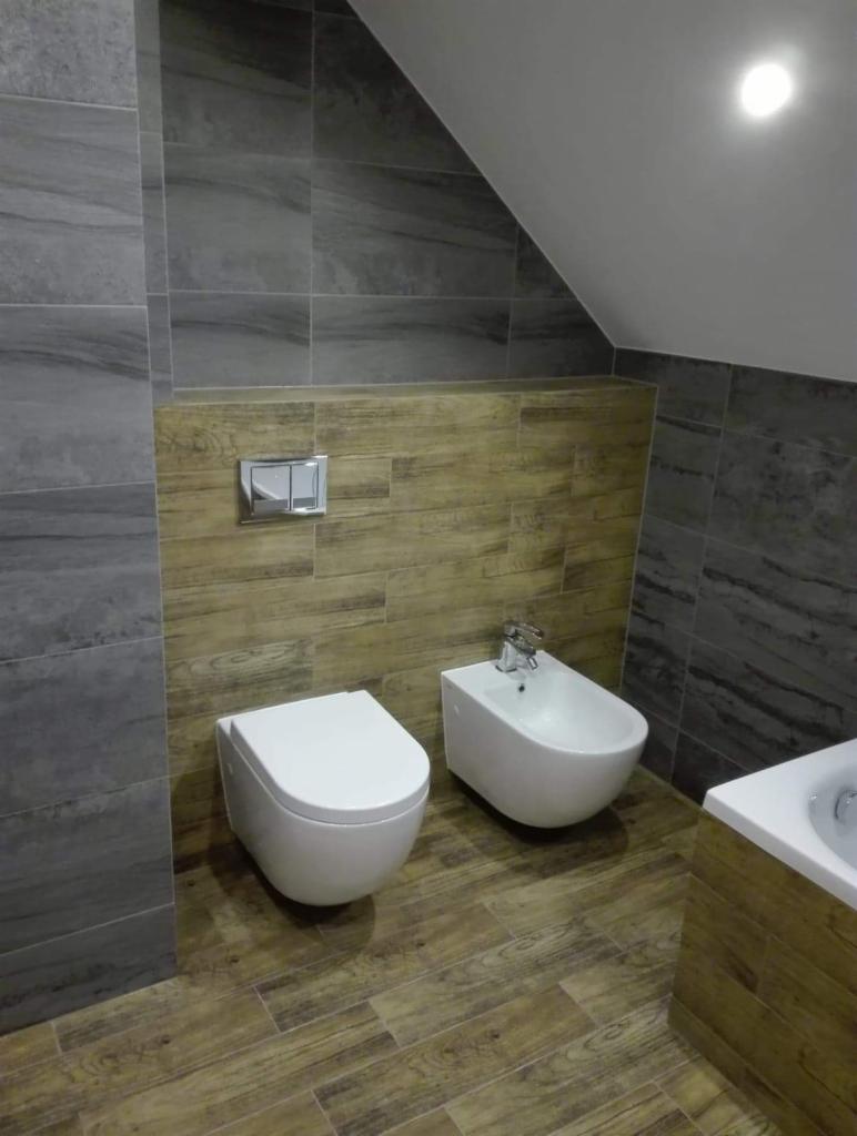 Nowoczesna łazienka z szarą i drewnopodobną glazurą, podwieszana toaleta i bidet w minimalistycznym stylu, widoczne elementy armatury i fragment wanny.