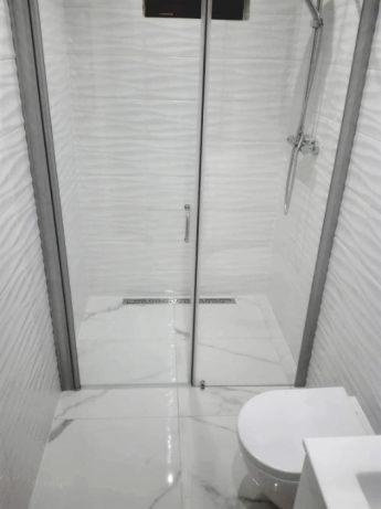 Nowoczesna kabina prysznicowa z białymi płytkami o falistej strukturze, szklane drzwi z metalowymi okuciami, odpływ liniowy w posadzce, biała ceramika łazienkowa.