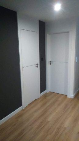 Dwa białe, minimalistyczne skrzydła drzwiowe z poziomymi frezowaniami, zamontowane w korytarzu z drewnopodobną podłogą i ścianami w kolorze grafitowym i szarym, oświetlone górnym światłem.