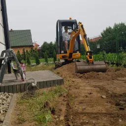 Operator w kabinie żółtej mini-koparki JCB, pracujący na placu budowy przy domu jednorodzinnym, widoczny fragment ułożonej kostki brukowej.