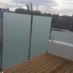 Szklana balustrada balkonowa z matowego szkła i nierdzewnych słupków, widok na zabudowę miejską w tle, drewniana podłoga balkonu.