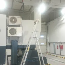 Dwa klimatyzatory Samsung zamontowane na ścianie obok urządzenia Nuctech w hali przemysłowej, drabina oparta o ścianę, widoczne elementy instalacji elektrycznej i przeciwpożarowej.
