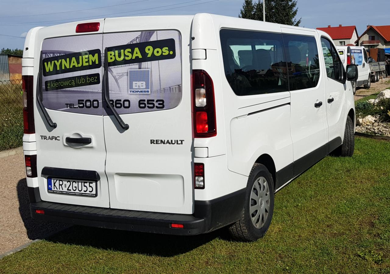 Tył białego busa Renault Trafic z reklamą wynajmu busa 9-osobowego z numerem telefonu, zaparkowanego na trawie.