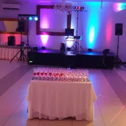 Sala bankietowa przygotowana na imprezę: scena z nagłośnieniem i oświetleniem, stół z kieliszkami wypełnionymi czerwonym płynem, dekoracje świetlne w kolorach niebieskim, różowym i fioletowym.