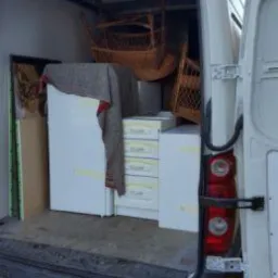 Wnętrze białego busa z otwartymi drzwiami, załadowane meblami: biała lodówka, komoda z szufladami i wiklinowe krzesła.