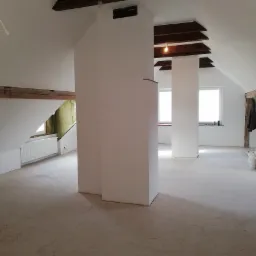 Poddasze po remoncie z drewnianymi belkami, oknami i świeżo pomalowanymi ścianami na biało, widoczna izolacja między belkami, podłoga przygotowana do dalszych prac wykończeniowych.