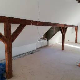 Poddasze w trakcie remontu, widoczne drewniane belki konstrukcyjne, częściowo wykończone ściany gipsowe, ocieplenie wełną mineralną oraz elementy instalacji elektrycznej.