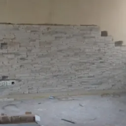 Fragment ściany z częściowo ułożoną dekoracyjną okładziną kamienną, widoczne gniazdka elektryczne, surowy stan wykończenia pomieszczenia.