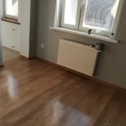 Pokój po remoncie: świeżo położone panele podłogowe o wzorze drewna, szare ściany i biały grzejnik pod oknem z drobnymi przedmiotami na parapecie.