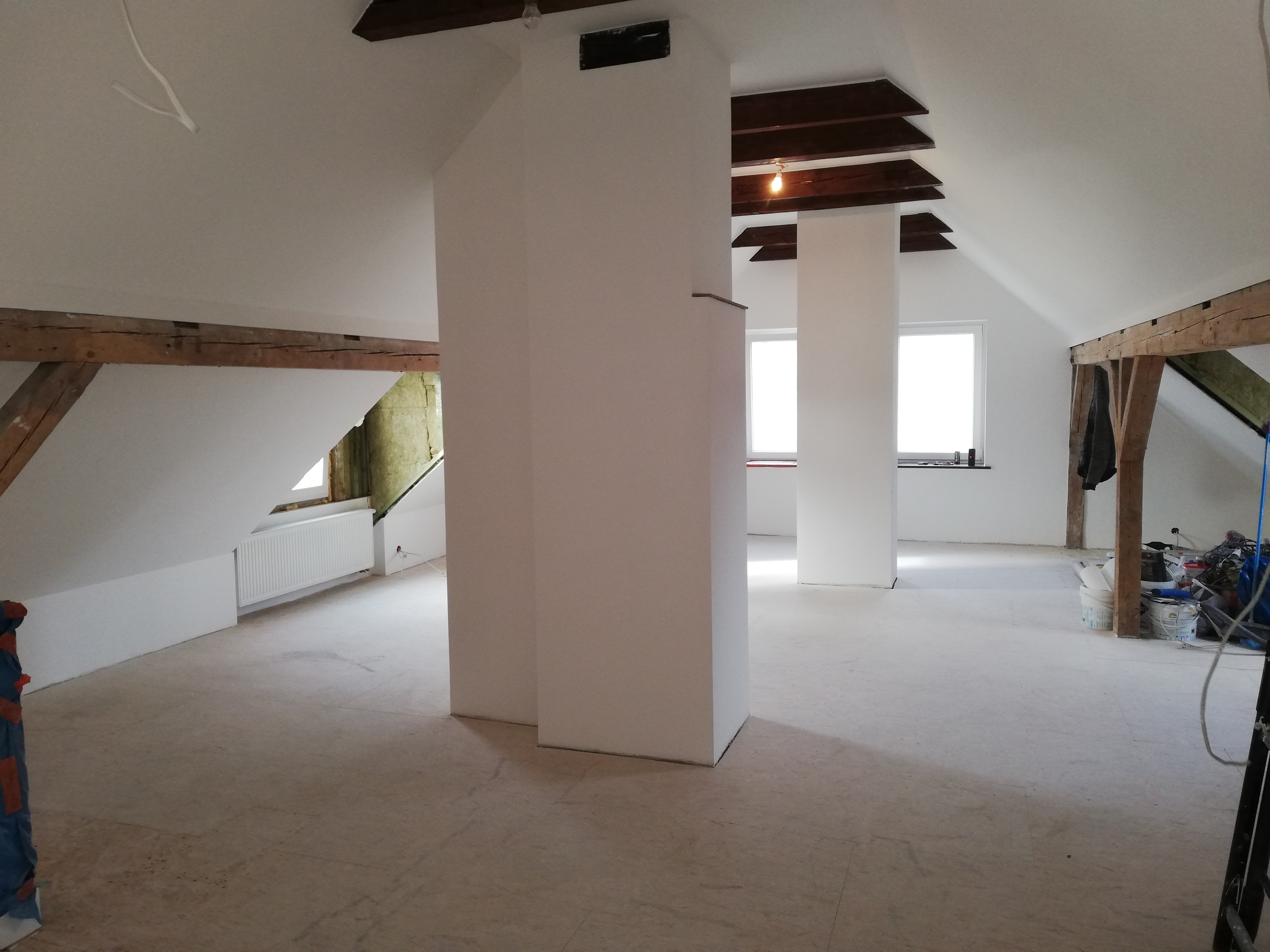 Poddasze po remoncie z drewnianymi belkami, oknami i świeżo pomalowanymi ścianami na biało, widoczna izolacja między belkami, podłoga przygotowana do dalszych prac wykończeniowych.