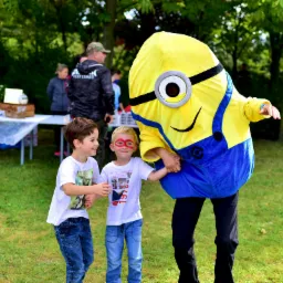 Dwaj chłopcy trzymają się za ręce, uśmiechając się do osoby przebranej za Minionka na zielonym trawniku podczas imprezy plenerowej.
