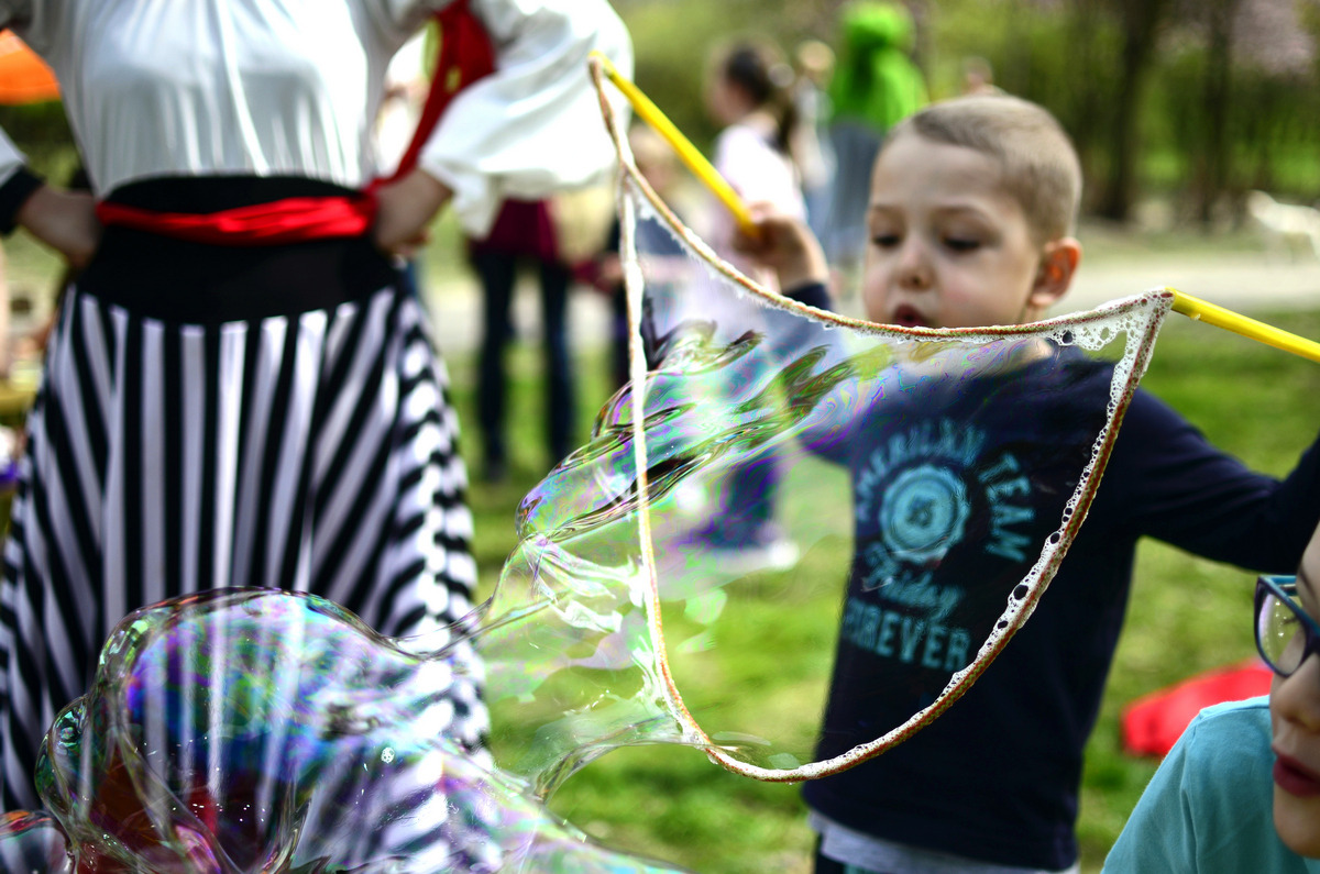Chłopiec trzymający kij z pętlą, tworzący bańki mydlane na tle osoby w stroju pirata i innych dzieci w parku.