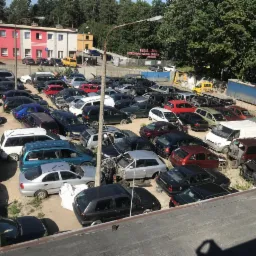 Widok z góry na zapełniony plac auto złomu z różnorodnymi samochodami, niektóre z widocznymi uszkodzeniami i brakującymi częściami, na tle budynku i drzew.