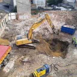 Żółta koparka głębi w wykopie na placu budowy, w tle gruz i fragmenty budynków, obok pracują robotnicy w kaskach.