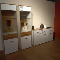 Zestaw mebli pokojowych w stylu nowoczesnym: dwie witryny z szklanymi półkami i szafka RTV z białymi frontami i drewnianymi akcentami, uzupełnione dekoracyjnymi wazonami i ramkami, prezentowane...