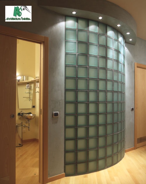 Zakrzywiona ściana z luksferów w szaro-beżowym wnętrzu, z widoczną łazienką w tle i punktowym oświetleniem sufitowym.