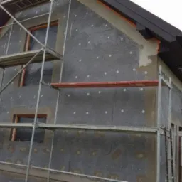 Dom jednorodzinny w trakcie prac związanych z montażem izolacji termicznej, widoczne rusztowania i płyty izolacyjne z kołkami mocującymi.