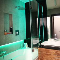 PROFES STUDIO Sylwia Fabjan-Jaros - Nowoczesna łazienka z wanną podświetloną turkusowym światłem LED, szklaną kabiną prysznicową i umywalką na drewnianej szafce z czarnym blatem.