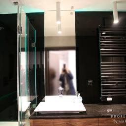 PROFES STUDIO Sylwia Fabjan-Jaros - Nowoczesna łazienka z czarną ścianą, podświetlanym lustrem i szklaną kabiną prysznicową, widok na umywalkę i grzejnik drabinkowy, odbicie osoby robiącej zdjęcie w lustrze.