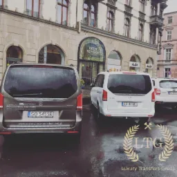 Dwa busy Mercedes-Benz V-klasa (brązowy i biały) zaparkowane przed hotelem Monopol na mokrym bruku, widoczne logo Luxury Transfers Group na pierwszym planie.