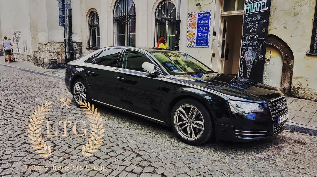 Czarna limuzyna Audi zaparkowana na brukowanej ulicy w europejskim mieście, z widocznym szyldem restauracji oferującej falafel i lody w tle. Na dachu pojazdu widoczna postać z jaskrawymi włosami.