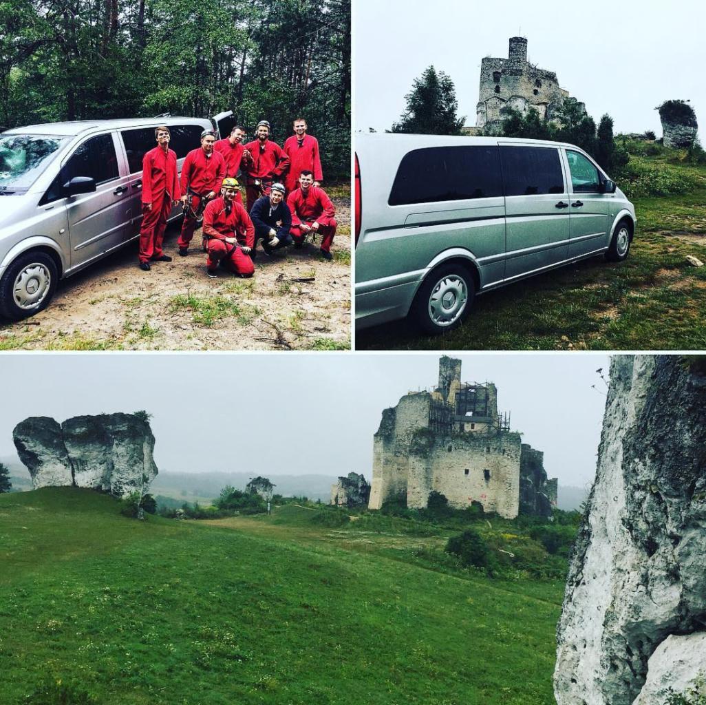 Grupa osób w czerwonych kombinezonach pozuje przy srebrnym vanie na tle leśnym i zamku w ruinie.