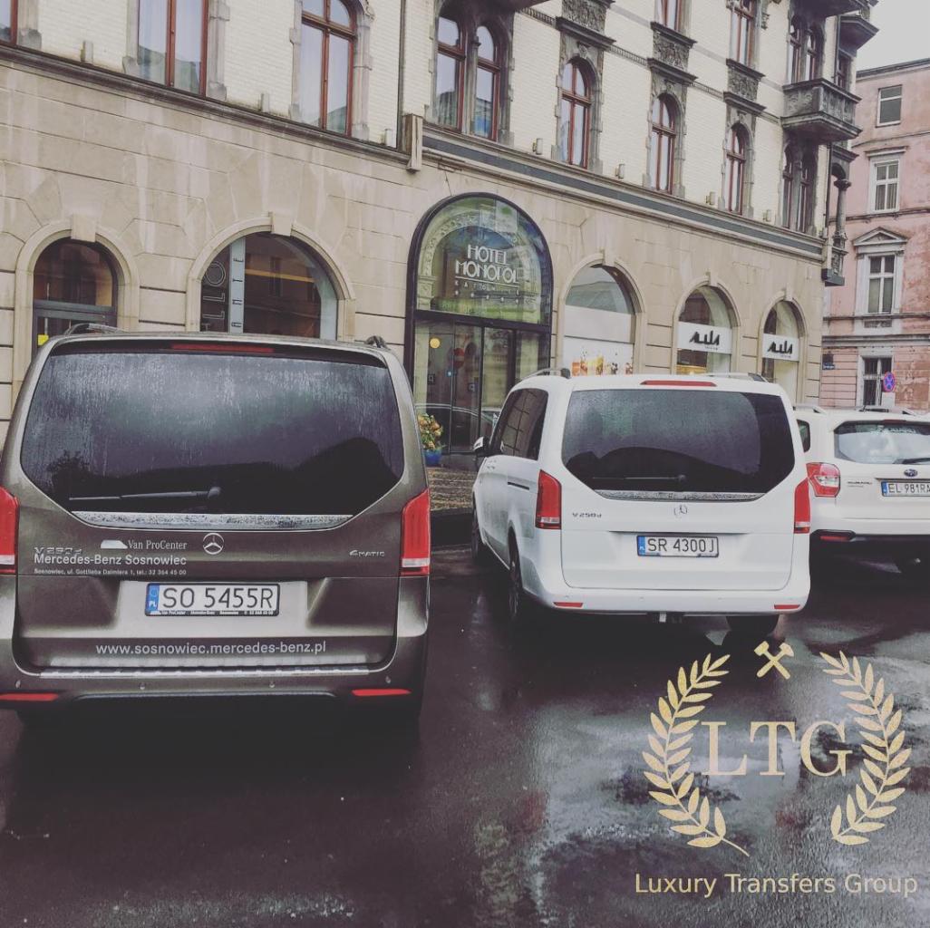 Dwa busy Mercedes-Benz V-klasa (brązowy i biały) zaparkowane przed hotelem Monopol na mokrym bruku, widoczne logo Luxury Transfers Group na pierwszym planie.