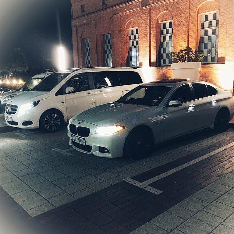 Dwa samochody zaparkowane nocą przed budynkiem z cegły: biały van Mercedes i srebrne BMW z czarnymi felgami.