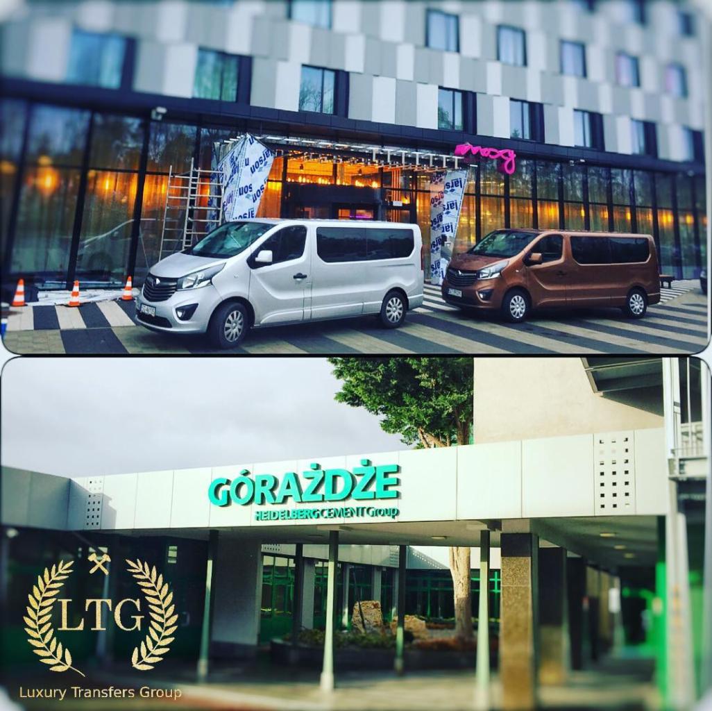 Dwa busy: srebrny i brązowy, zaparkowane przed budynkiem z napisem 'Larson' i 'Roxxy', w tle widoczne rusztowania i stożki ostrzegawcze. W dolnej części widać budynek z napisem 'Górażdże' i logo...