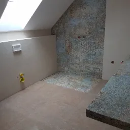 Łazienka na poddaszu w trakcie układania płytek: beżowe płytki podłogowe i ścienne, dekoracyjna ściana z płytek wzorzystych, żółte rury instalacyjne, okno dachowe.