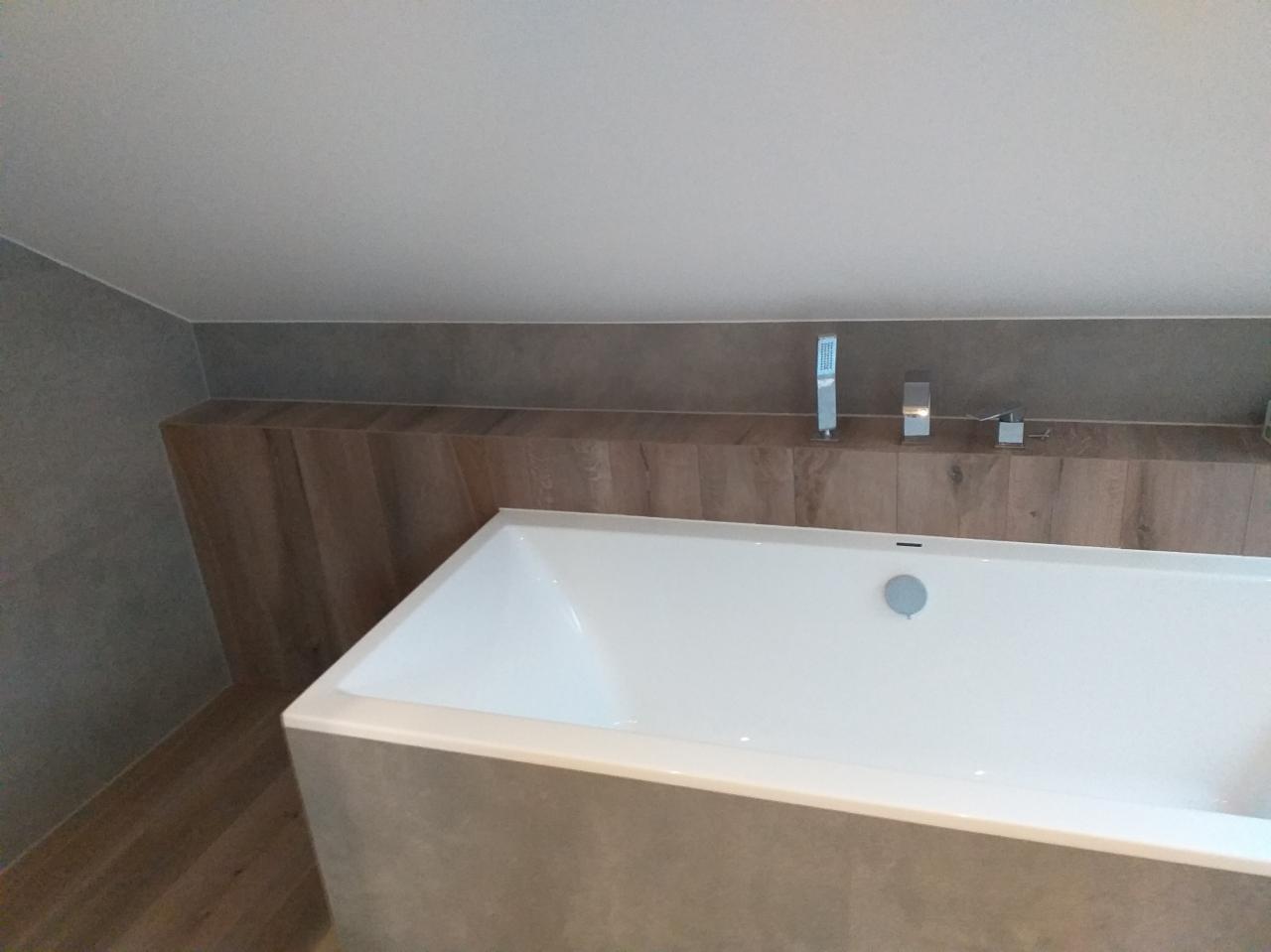 Wanna w obudowie z szarych płytek, drewnopodobna wnęka z armaturą, minimalistyczne wykończenie łazienki na poddaszu.