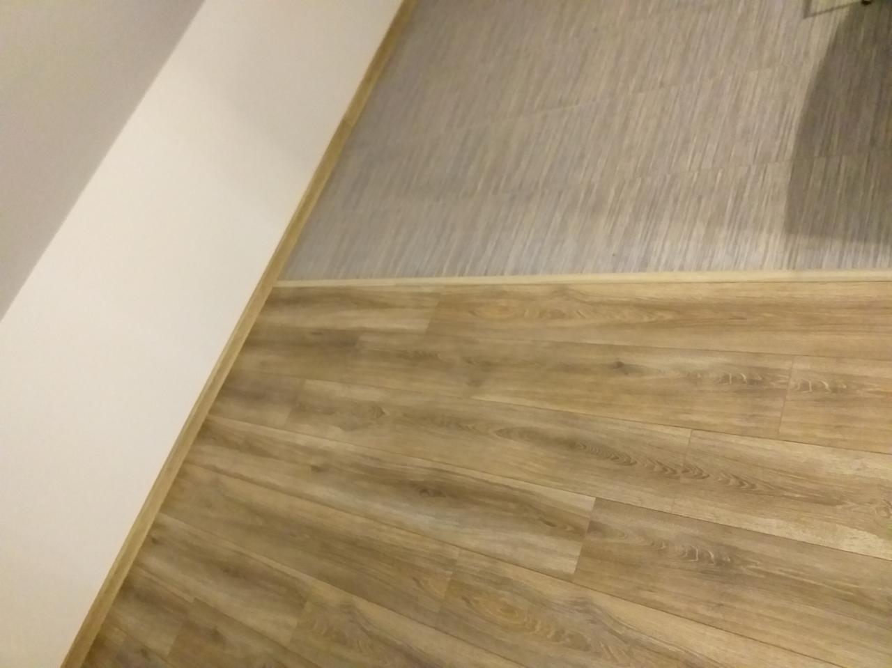 Połączenie paneli podłogowych imitujących drewno z szarą tapetą strukturalną na ścianie, listwy przypodłogowe w kolorze podłogi, realizacja w Żabnie.