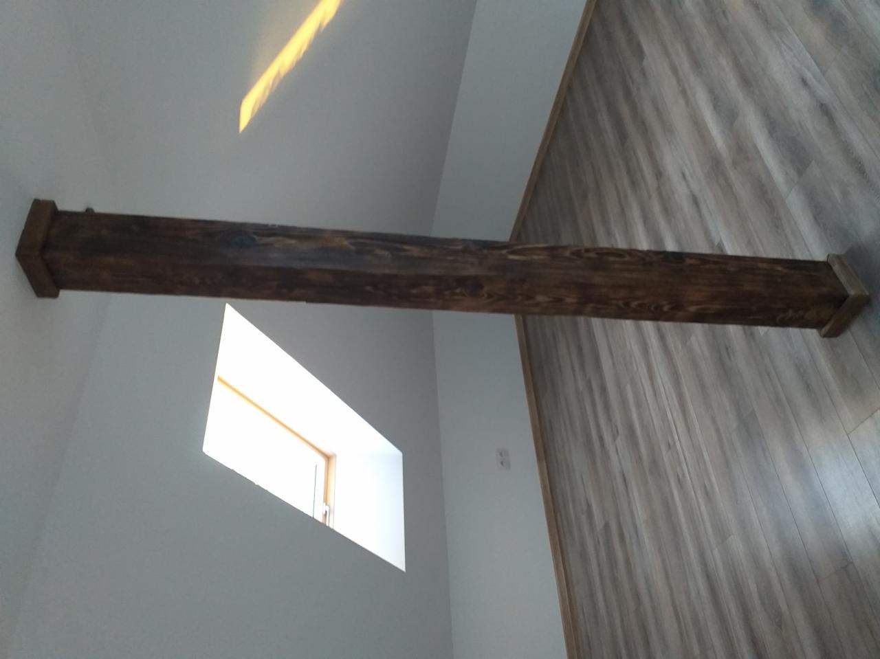 Rustykalna belka stropowa z ciemnego drewna, zamontowana w jasnym wnętrzu z drewnopodobną okładziną ściany i oknem w tle.