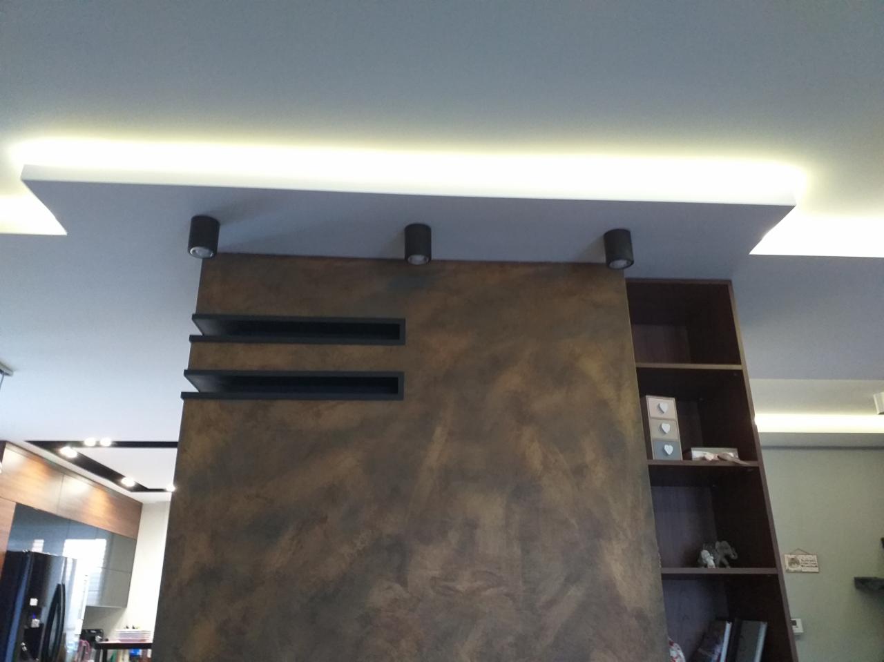 Ściana z dekoracyjnym tynkiem w odcieniach brązu, z wnękami w formie półek i oświetleniem punktowym, sąsiadująca z regałem na książki i elementami zabudowy kuchennej w tle.