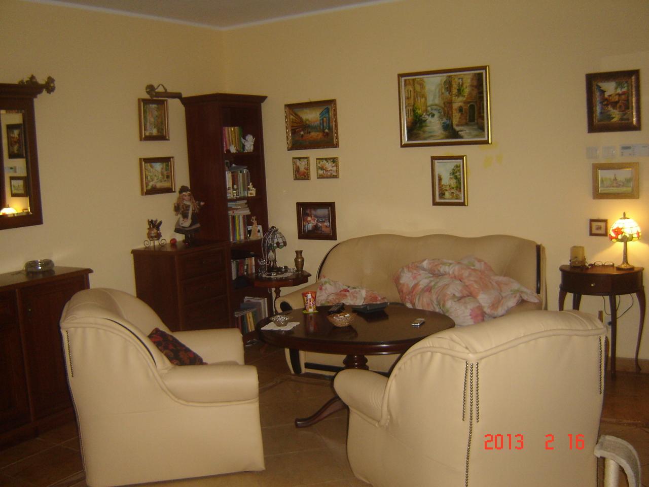 Salon w Rokietnicy z beżowymi ścianami, ciemnymi meblami, skórzanym fotelem z ozdobnymi nitami, okrągłym stolikiem kawowym i licznymi obrazami na ścianach.