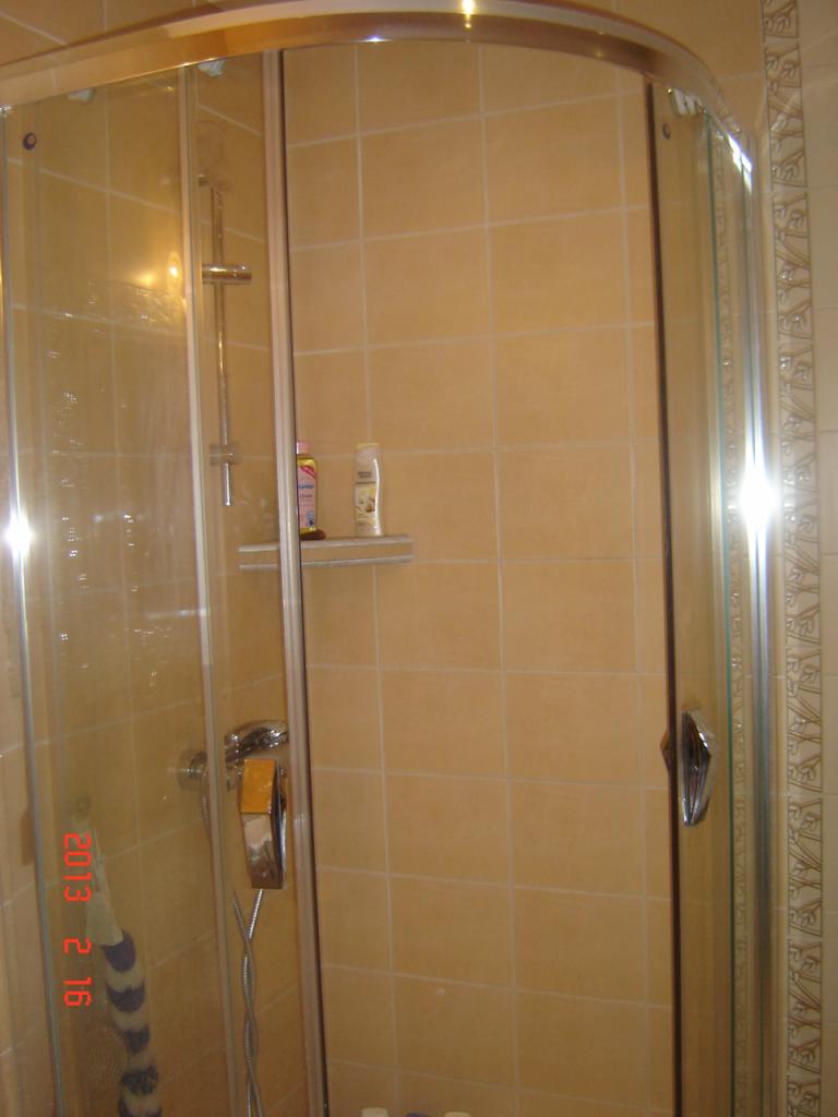 Wnętrze łazienki z kabiną prysznicową z zaokrąglonymi szybami, wyłożonej beżowymi płytkami ceramicznymi, z widocznymi kosmetykami na półce i datą 2013 2 16 odbitą na szybie.