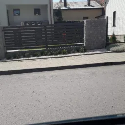 Nowoczesne ogrodzenie z poziomych paneli w kolorze ciemnego brązu, osadzone na kamiennych słupkach, z niską roślinnością i białym grysem u podstawy, widoczne z ulicy.