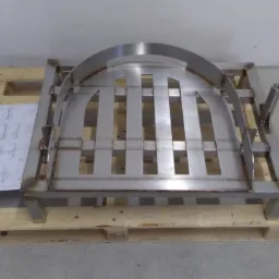 Metalowa konstrukcja z elementami giętymi i spawanymi, przypominająca bramę lub fragment pieca, umieszczona na drewnianej palecie obok rysunku technicznego.