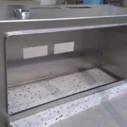 Metalowa obudowa urządzenia, wykonana z polerowanej stali nierdzewnej, z widocznymi otworami i spawami, ustawiona na szarym, nakrapianym cokole.