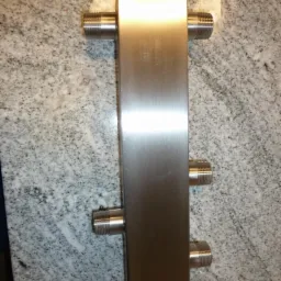 Pionowo ułożony, srebrny, prostokątny wymiennik ciepła z sześcioma gwintowanymi króćcami na tle szarego blatu z granitu.