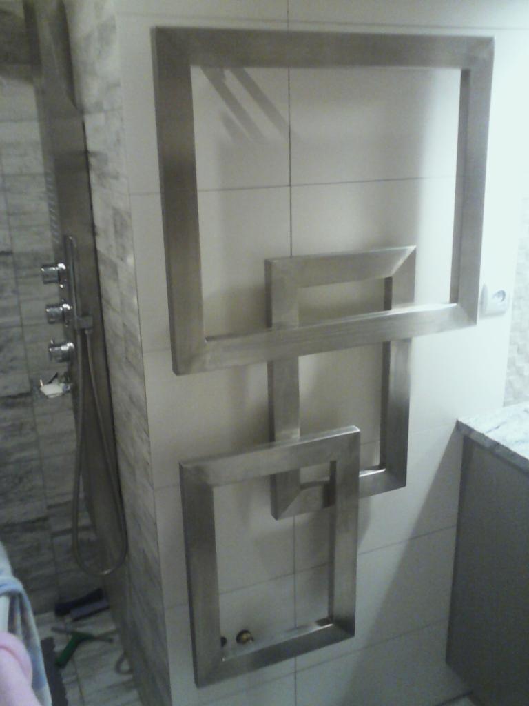 Metalowa dekoracja ścienna w łazience, składająca się z połączonych, kwadratowych ram z metalu, zamontowanych na białych płytkach.