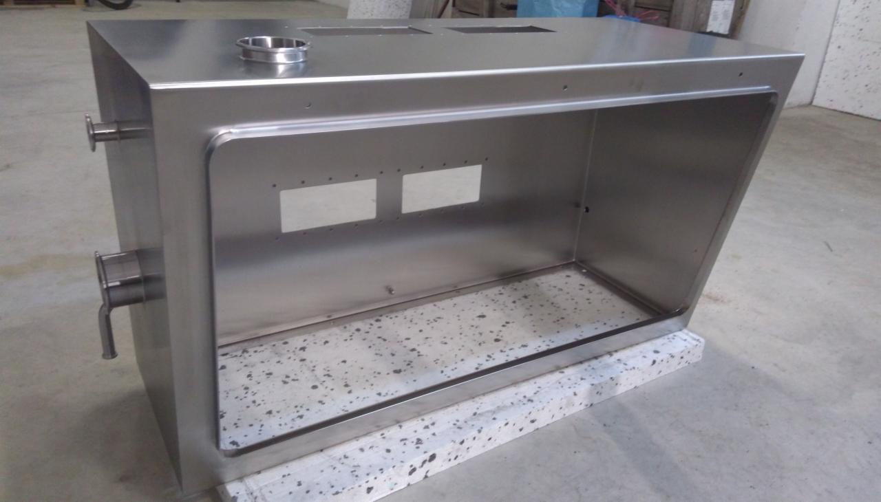 Metalowa obudowa urządzenia, wykonana z polerowanej stali nierdzewnej, z widocznymi otworami i spawami, ustawiona na szarym, nakrapianym cokole.