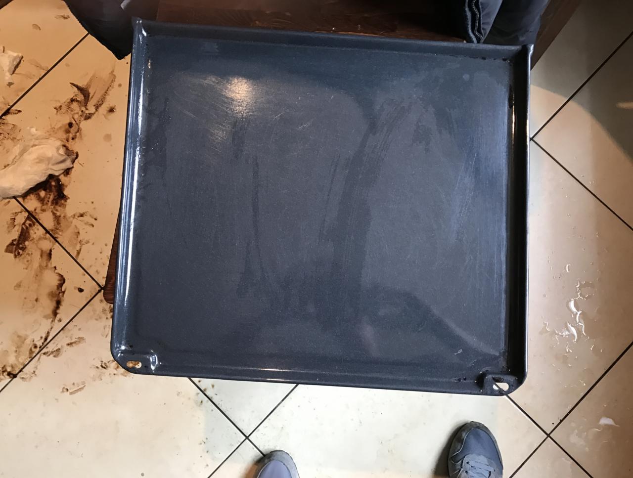 Blacha piekarnika po czyszczeniu, widoczne ślady po myciu, na podłodze zabrudzenia i ślady wody, fragment buta sportowego w rogu kadru.