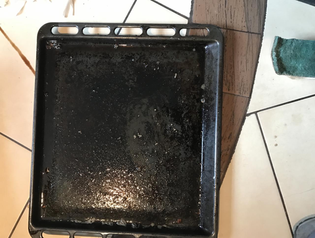 Brudna, tłusta blacha do pieczenia leżąca na podłodze z płytek, obok zielonej gąbki. Widoczne resztki przypalonego jedzenia i tłuszczu.
