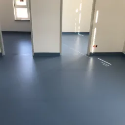 Niebieska, gładka podłoga z listwami przypodłogowymi w nowym, pustym pomieszczeniu z białymi ścianami i otwartymi drzwiami, wpadające promienie słoneczne.