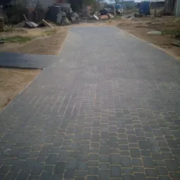 Ułożona szara kostka brukowa na podjeździe, widoczne piaskowe spoiny i nierówności terenu w tle.