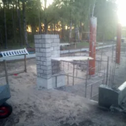 Wznoszenie fundamentów pod budynek z widoczną konstrukcją zbrojeniową, fragmentem ściany z pustaków oraz taczką na pierwszym planie, w tle zalesiony teren.