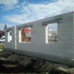 Wznoszenie ścian parteru z białych bloczków betonu komórkowego. Widoczne otwory okienne z nadprożami. Na pierwszym planie wykopany grunt pod fundament.